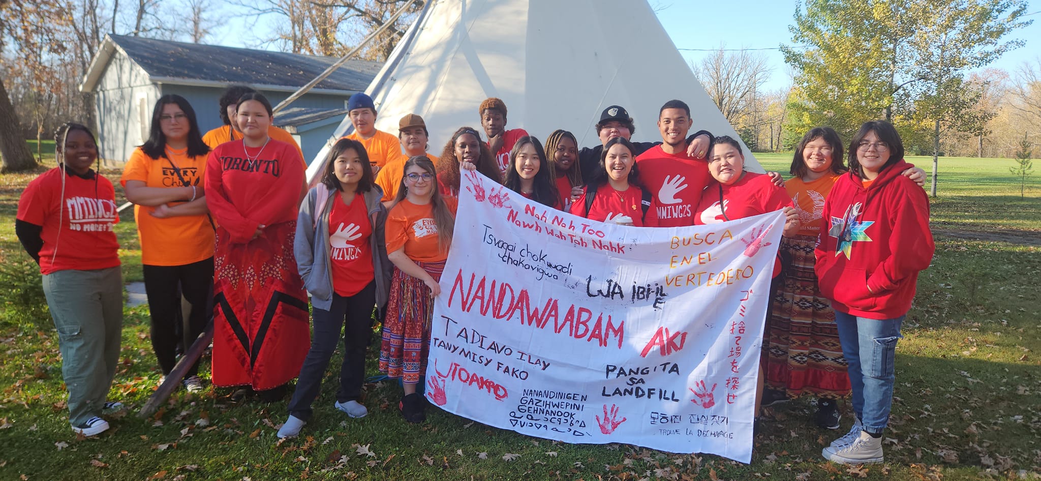 Indigenous and racialized youth hold a banner saying "Search the Landfill" in many languages.