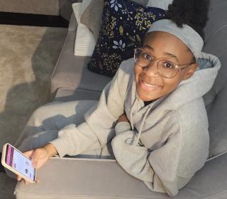 Jonisha Lewinson, a teenage girl dressed in a grey hoodie, sits on a couch holding her smartphone 