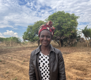 A woman wearing an African headscarf stands smiling against a background of a field with tilled rows.