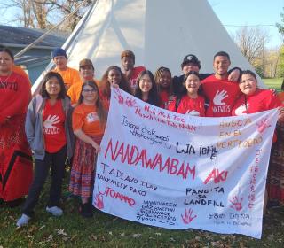 Indigenous and racialized youth hold a banner saying "Search the Landfill" in many languages.