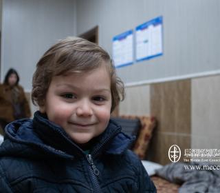 A small boy in a dark blue jacket smiles in a room with mattress on the floor.