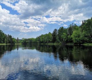 Sunny day on a lake surrounded by trees, the clouds reflecting in the water.