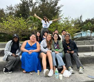 Eleven young people sit on steps smiling and waving at the camera