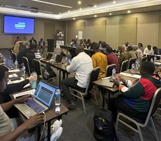 A packed conference room lined with people writing or on laptops. Four presenters sit at the front.