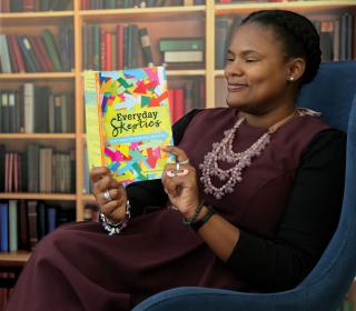 Lenten book editor Alydia Smith, dressed in a long purple dress and detailed necklace, sits in a large comfy to read a copy of the book "Everyday Skeptics."