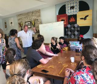 The group of GC43 Pilgrims makes dreamweavers in the office of the All Native Circle Conference. A large quilt symbolizing the Four Directions appears on the wall behind the group.