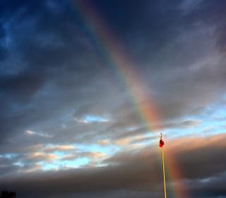 A photo of a Canadian flag on a flagpole apparently being found at the foot of a rainbow, against the contrast of a dramatic dark sky.