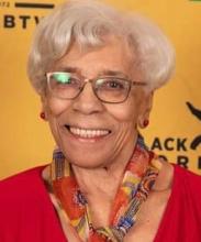 A portrait of author Nancy Oliver-MacKenzie, a Black elder wearing a red top and multicolour scarf as she smiling joyously for the camera.