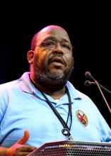 Paul Douglas Walfall, a Black man with glasses and a graying beard, in a blue shirt, is shown from the shoulders up, speaking to General Council.