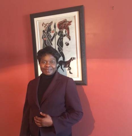 The Rev. Marie-Claude Manga, standing in front of an artwork, dressed in a brown jacket.