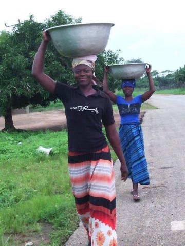 Two Zambian women walk along a road carrying large metal tubs on their heads.