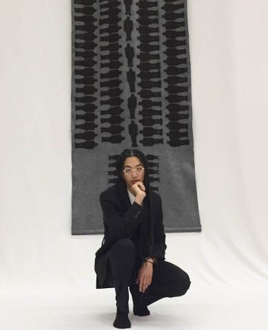 A young Black man in a black suit with locks and golden glasses, poses in front of one of his art works, a depiction of a trans-Atlantic slave ship holdings.