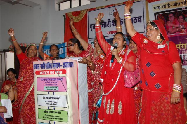 Seven Indian women dressed in red stand at a podium with their right arms raised. 