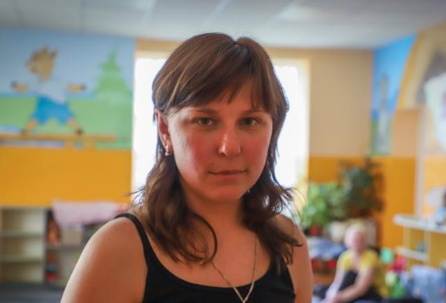 A woman in her 30s looks into the camera. In the background we see a room decorated with children's art and toys.