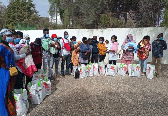 Several people, many women holding babies, are lined up outdoors and have the same bag of food in front of each of them.