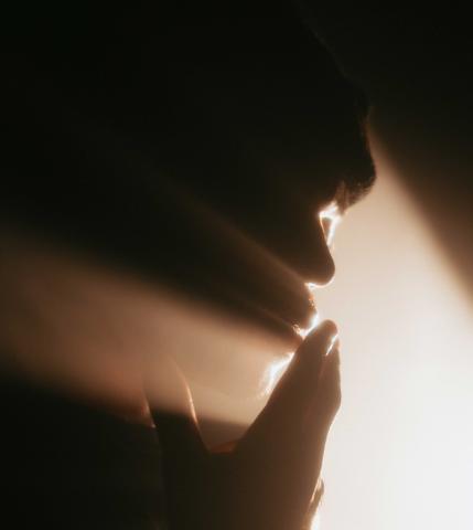 A silhouette of a person touching their lips in prayer, as light pours between their fingers.