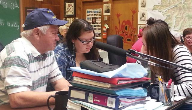 An older man and two young women sit at a desk stacked with papers, books, and binders. In the background are more small groups sitting at tables. 