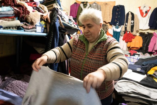 A woman with short white hair holds up clothes while folding them, in a store room of supplies for Ukraine refugees.