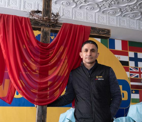 A man with dark hair and a compassionate face, looks into the camera as he stands on an altar beneath a cross decorated with a red cloth and crown of thorns.