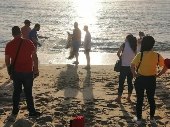 Two men prepare to cast a fishing net into the ocean as a several people on the beach look on.