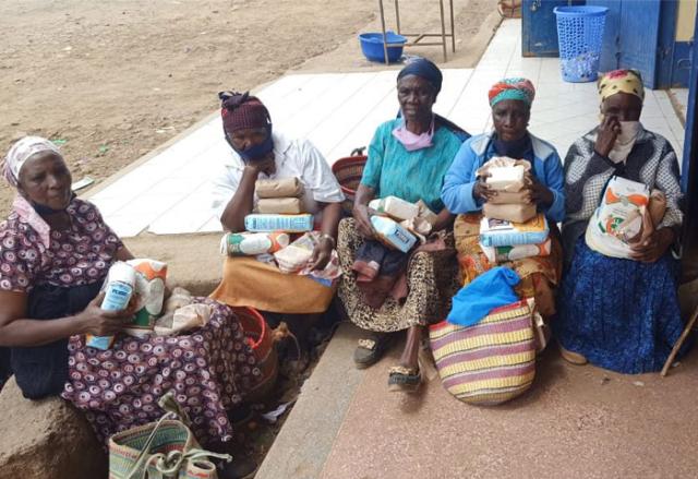 Five older African women are seated on the porch of a building holding parcels and shopping bags.