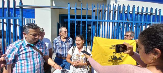 Group of people standing outside a building. A woman standing in the centre is speaking while another woman holds her phone up to record her.