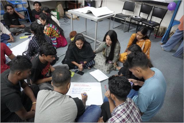 Several young people sit in a circle on the floor of a classroom making notes and talking.