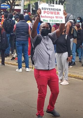 A man wearing a clerical collar holds up a sign that says "The sovereign power belongs with the people." 