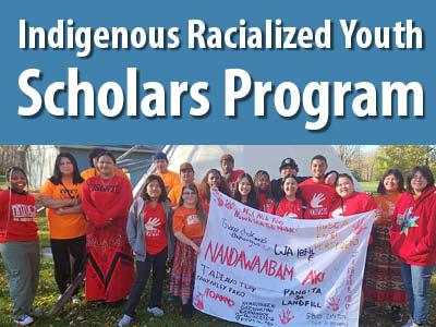 A large group of young people in orange shirts stand in front of a tepee hold a banner with Indigenous words on it