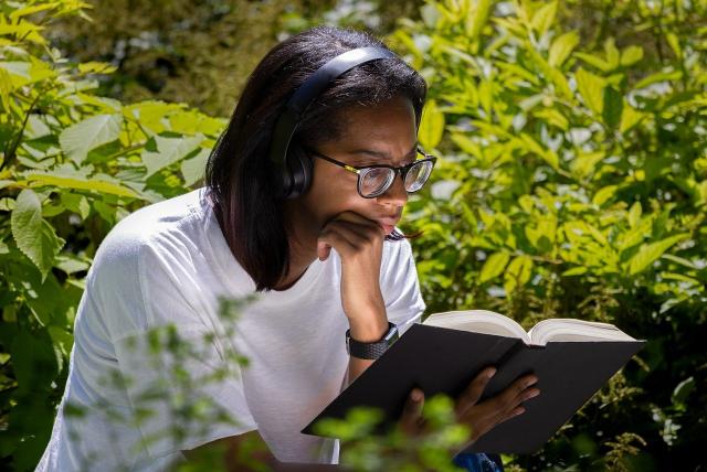 Young woman in white shirt reads a book outdoors, surrounded by trees.