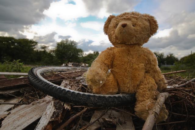 A discarded orange teddy bear sits atop an old bicycle tire and other garbage in a dump.