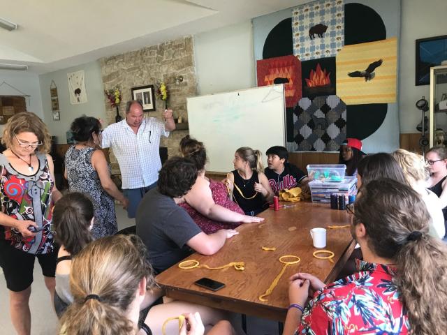 The group of GC43 Pilgrims makes dreamweavers in the office of the All Native Circle Conference. A large quilt symbolizing the Four Directions appears on the wall behind the group.