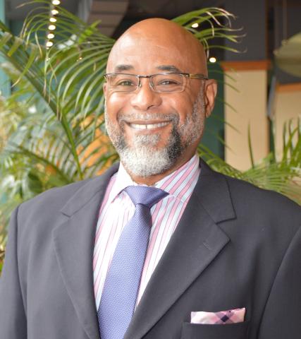 A portrait of Rev. Dr. Bentley de Bardelaben-Phillips, a Black man with glasses and a thin black and grey beard. He has a bright smile and is wearing a dark suit.