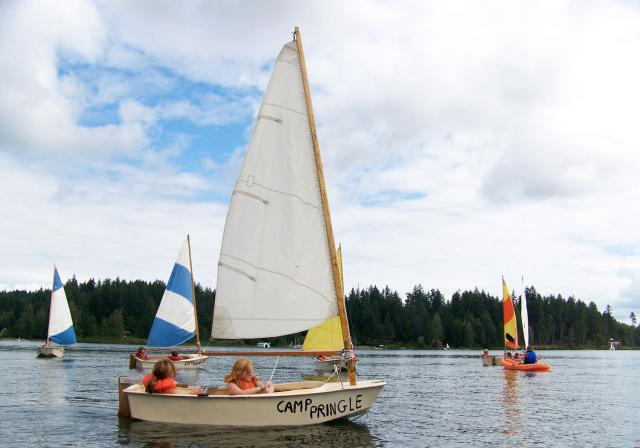 A small sailboat being piloted by two children in lifejackets in a lovely lake surrounded by forest. In the background are a number of similar boats piloted by camp kids.