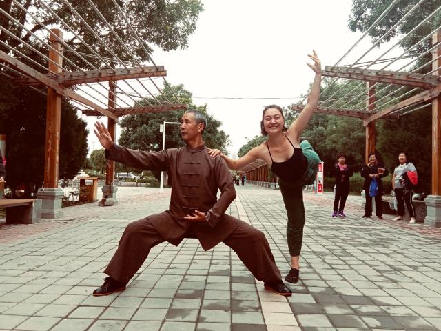 The author, a young White woman, balances in a ballet pose holding the shoulder of an elderly Tai Chi master in the streets of Jiuquan, China while amused people look on.