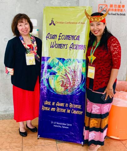 Author Kim Uyede-Kai from the United Church stands with Rev. Ester Damaris Wolla Wunga from Indonesia stand with an assembly banner. Both women are of Asian background, but the "traditional dresses" they are wearing reflect their different cultures.