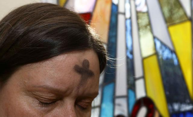 A White woman with Lenten ashes on her forehead is shown from the eyes up, and stands near a colourful stained glass window.