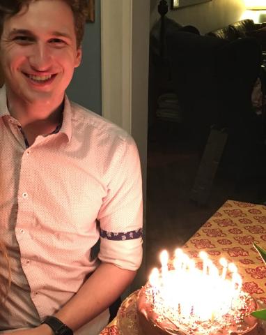 A young white man standing in a door frame, illuminated by the candles on the cake for his birthday.