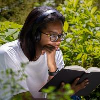 Young woman in white shirt reads a book outdoors, surrounded by trees.