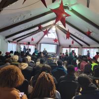 Young people fill a large room watching a presentation at the front.